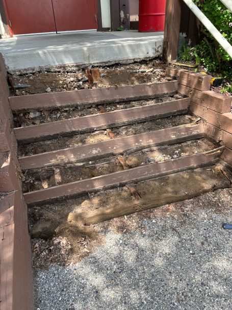 Deteriorated wooden steps with uneven surfaces and exposed soil beside a building entrance.